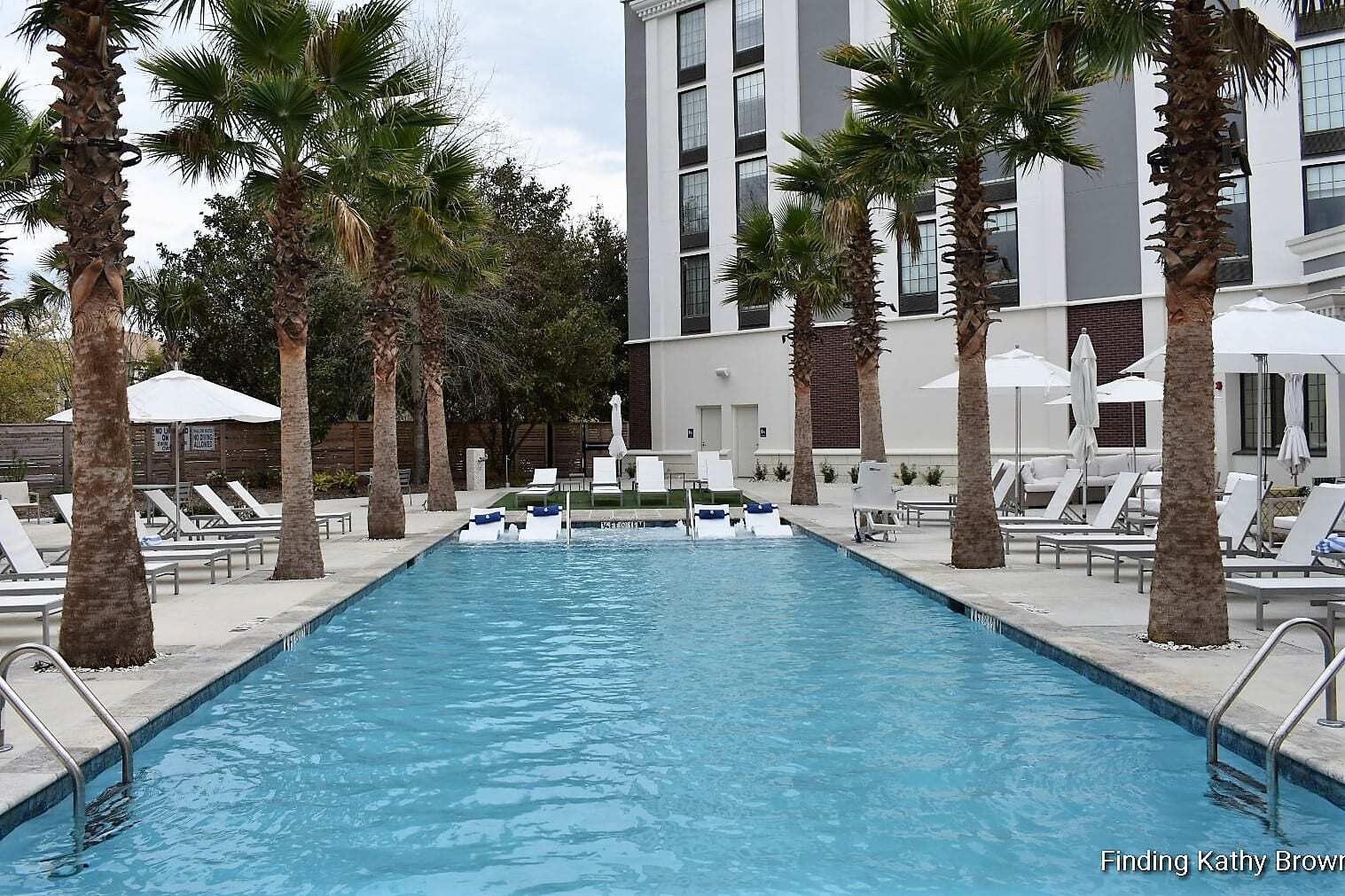 "Relaxing Resort-Style Pool at Hotel Indigo Mt Pleasant, South Carolina. Enjoy a serene oasis with palm trees, umbrellas, and comfortable loungers. Sunk-in lounge chairs in the shallow end offer the perfect spot for ultimate relaxation."