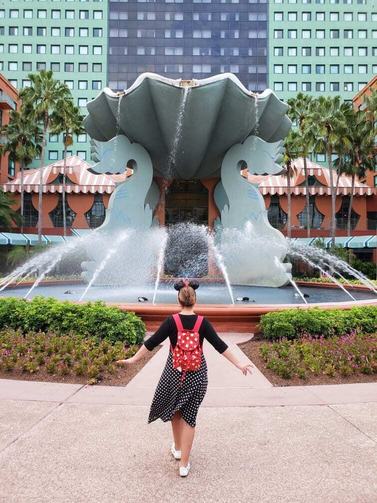 "Chattanooga travel blogger Kathy Brown, dressed in a stylish black and white polka dot dress, wearing vibrant red sequined Minnie Mouse ears and carrying a sequined backpack, strolls towards a magnificent Swan fountain at the Swan and Dolphin Resort in Orlando, Florida. With a sense of adventure in her stride, Kathy explores the enchanting surroundings, capturing the essence of Disney magic. The Swan fountain stands tall, its graceful beauty mirroring Kathy's excitement and joy during her visit. Join Kathy on her magical journey at the Swan and Dolphin Resort."