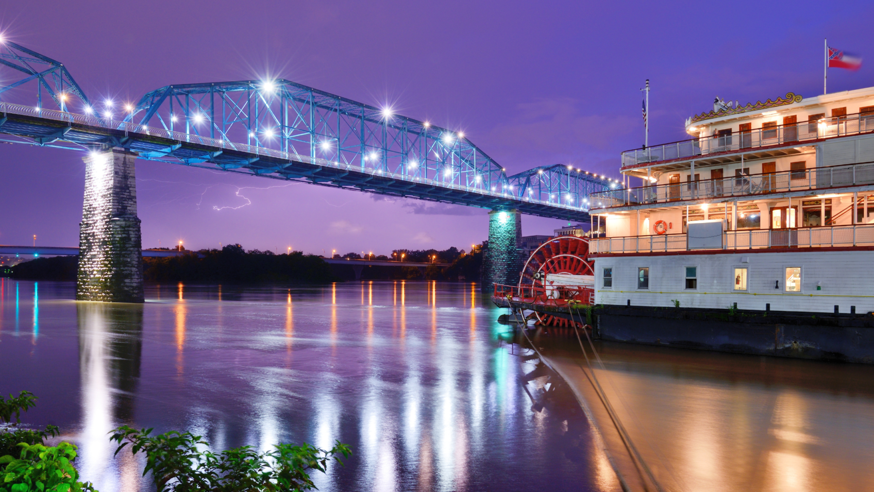 "Tennessee River in Chattanooga, TN at night with a view of a bridge illuminated by twinkling lights in the background. The Southern Belle dinner cruise sailing under the bridge adds to the scenic view."