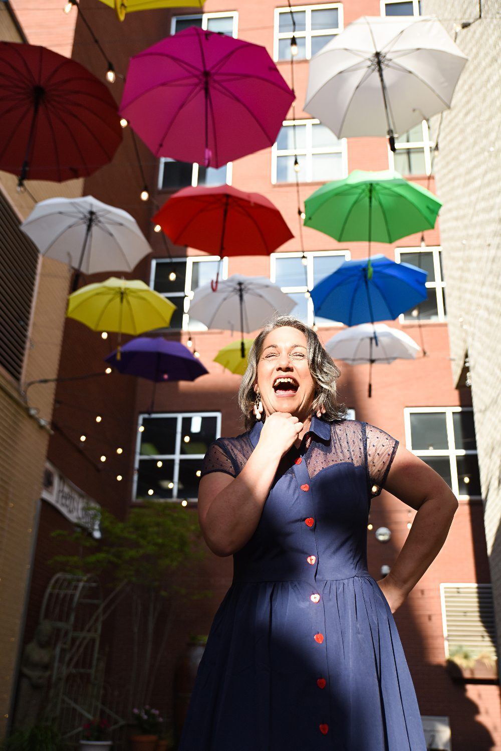"Chattanooga travel blogger Kathy Brown stands under a colorful umbrella canopy in a hip alley, wearing a blue retro dress with pink buttons and smiling with excitement in her eyes."