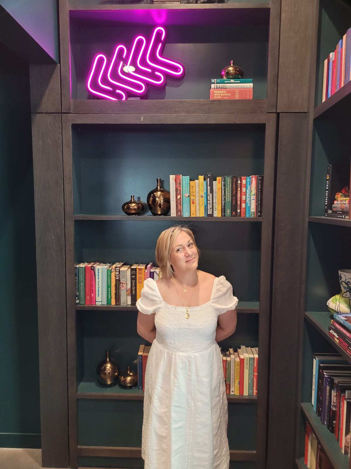 Blonde haired woman stands smiling in front of a bookcase. Books fill the floor to ceiling bookcase and 4 pink neon lights point downward.