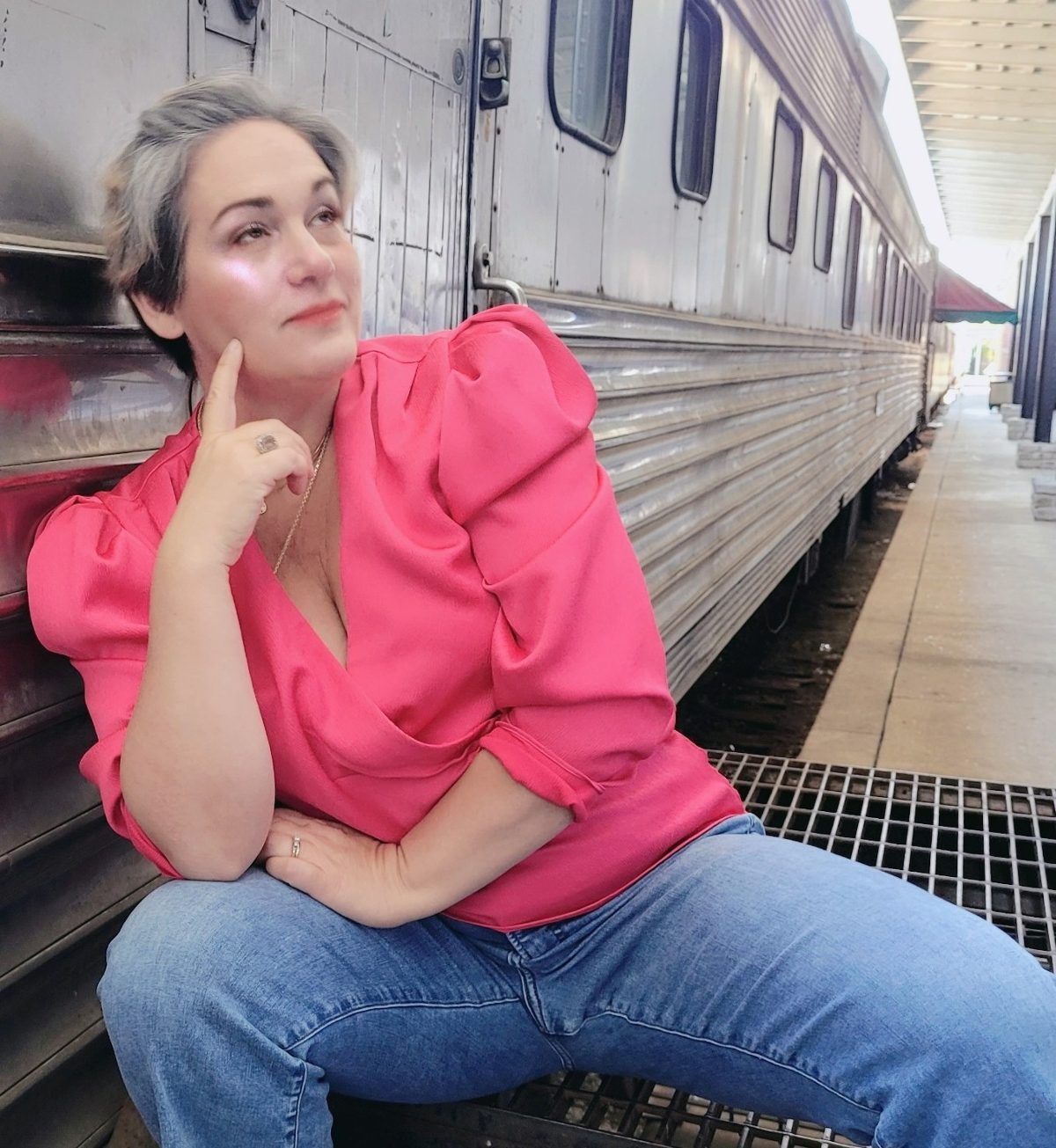 Going Grey on Purpose "Kathy Brown, a top Travel Blogger in Chattanooga, is wearing a pink blouse and casually looking off into the distance. Her short, gray hair reflects her age-positive influence. She is kneeling against the Chattanooga Choo Choo."
