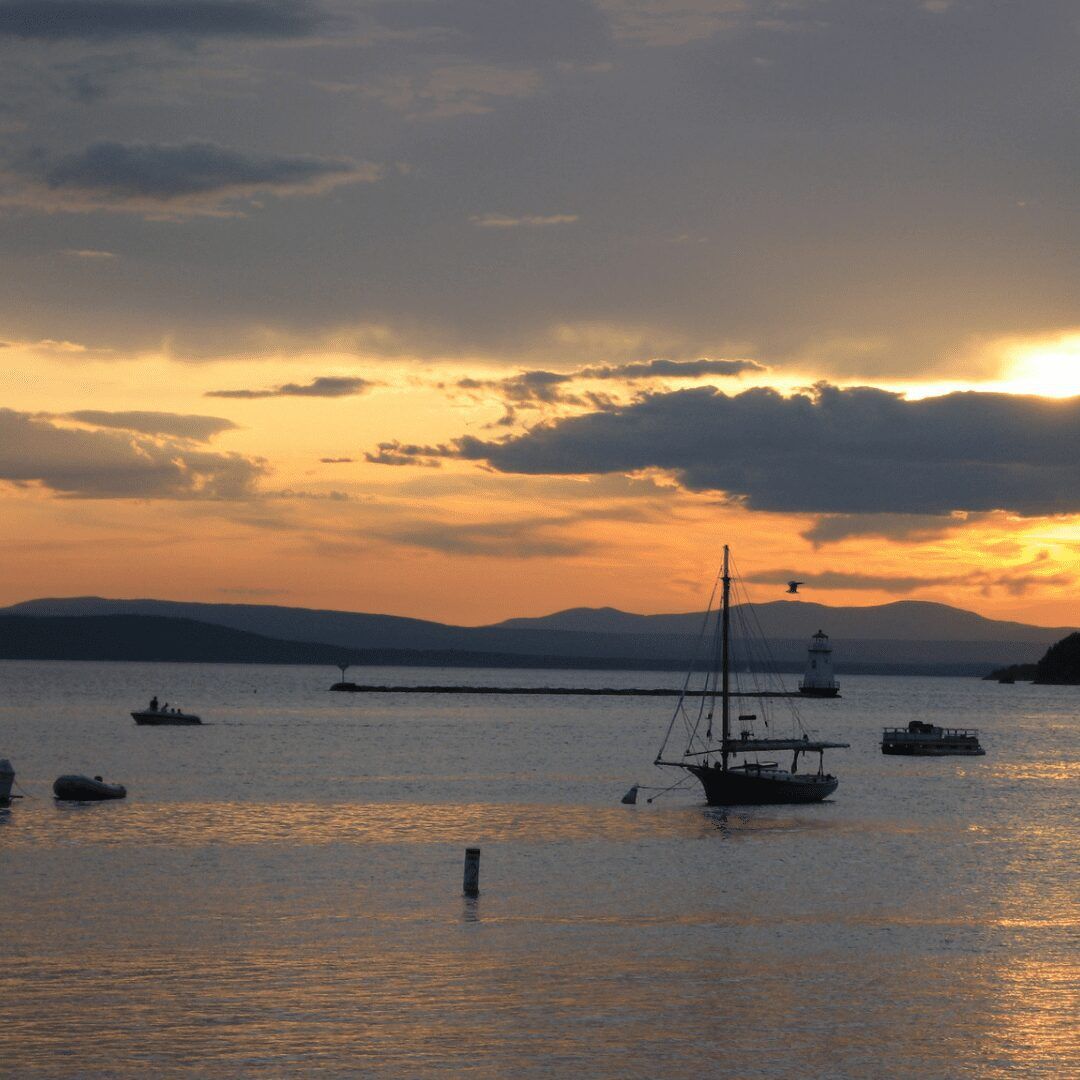 An image of Lake Champlain in Vermont with crystal-clear blue water and a mountainous landscape in the background. The sun is shining brightly, casting a warm glow on the tranquil scene. A few boats are visible in the distance, adding to the peaceful and serene atmosphere of the area."