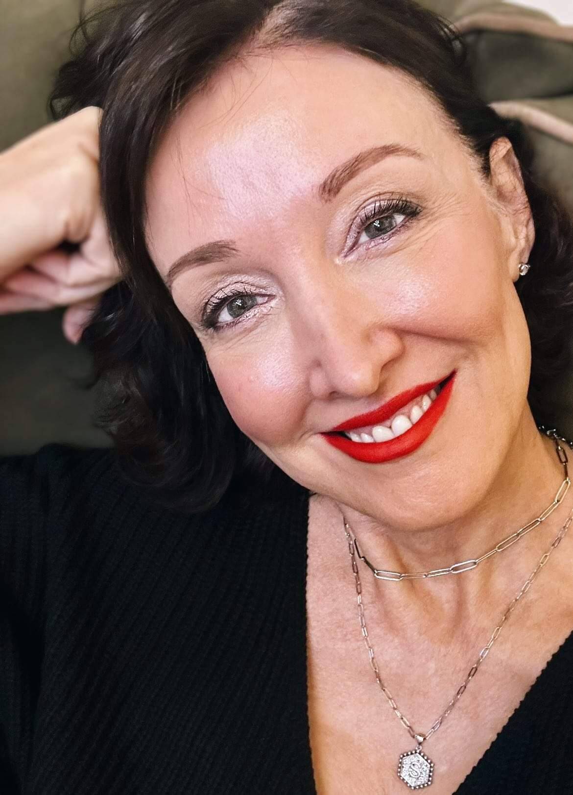 Suzan Hall, an over 50 GenX enthusiast, with short black hair, a slender build, and impeccable makeup, including striking red lipstick. She's proudly sporting a GenX t-shirt, radiating confidence and style.