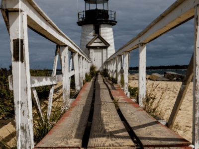 "View of a white lighthouse with a black roof on Nantucket Island, Massachusetts, from the perspective of walking over a wooden boardwalk. The boardwalk leads up to the lighthouse, which overlooks the sea and is surrounded by greenery and sandy dunes."
