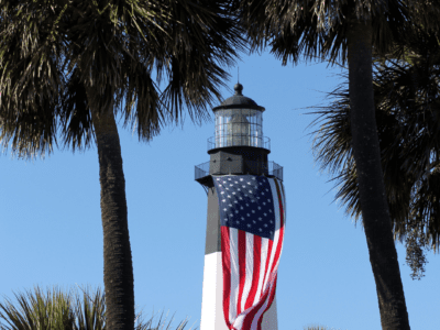 The Tybee Lighthouse stands tall against the blue sky, with the American flag waving in the wind. The white and black striped tower is topped with a red roof and surrounded by greenery.