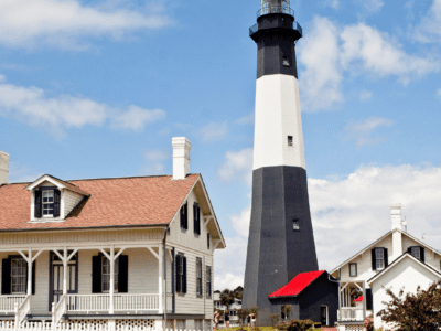 "Tybee Island Lighthouse - a towering white and black striped lighthouse with red rooftops, surrounded by green trees and blue sky."