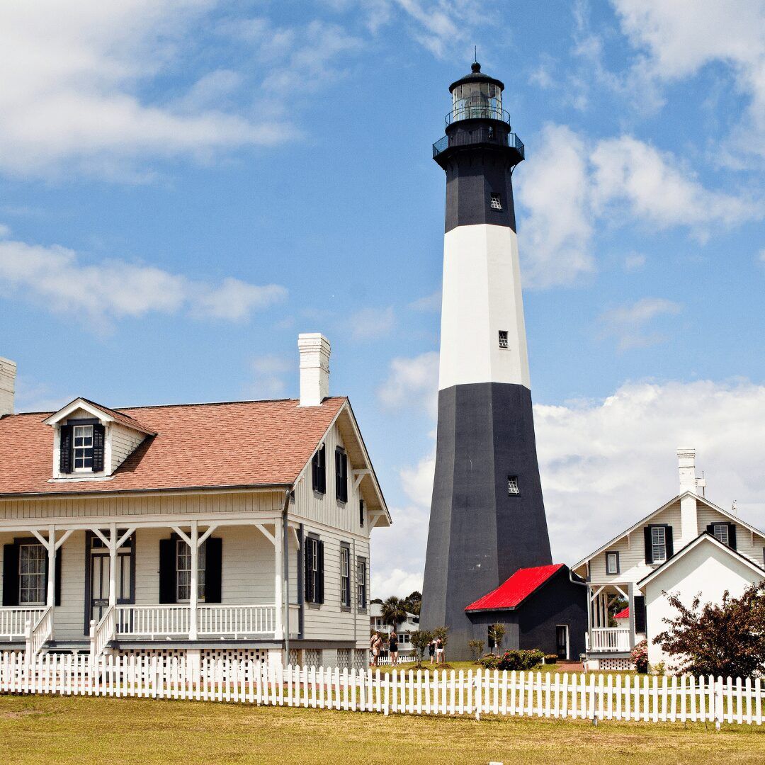"Tybee Island Lighthouse - a towering white and black striped lighthouse with red rooftops, surrounded by green trees and blue sky."