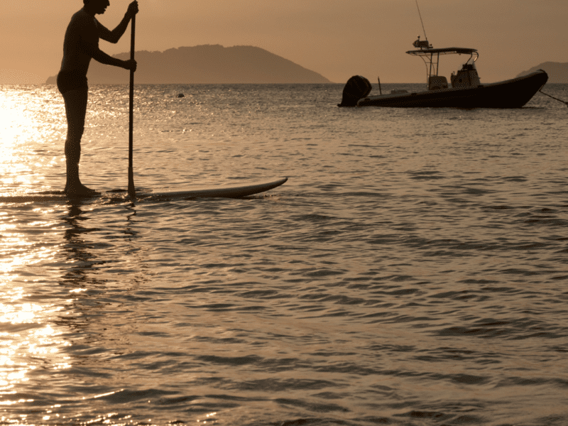 sunset view of a man on a paddle-board in the middle of a lake. Behind him is a fishing boat.