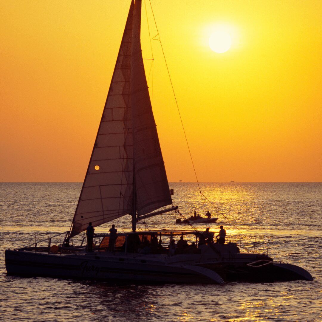 "An orange and pink-hued sunset sky fills the background, with a sailboat gliding across calm, turquoise waters in Key West, Florida. The boat's white sails are catching a warm, gentle breeze as it cuts through the water, leaving a small trail of ripples in its wake. Palm trees dot the shoreline in the distance, silhouetted against the colorful sky."