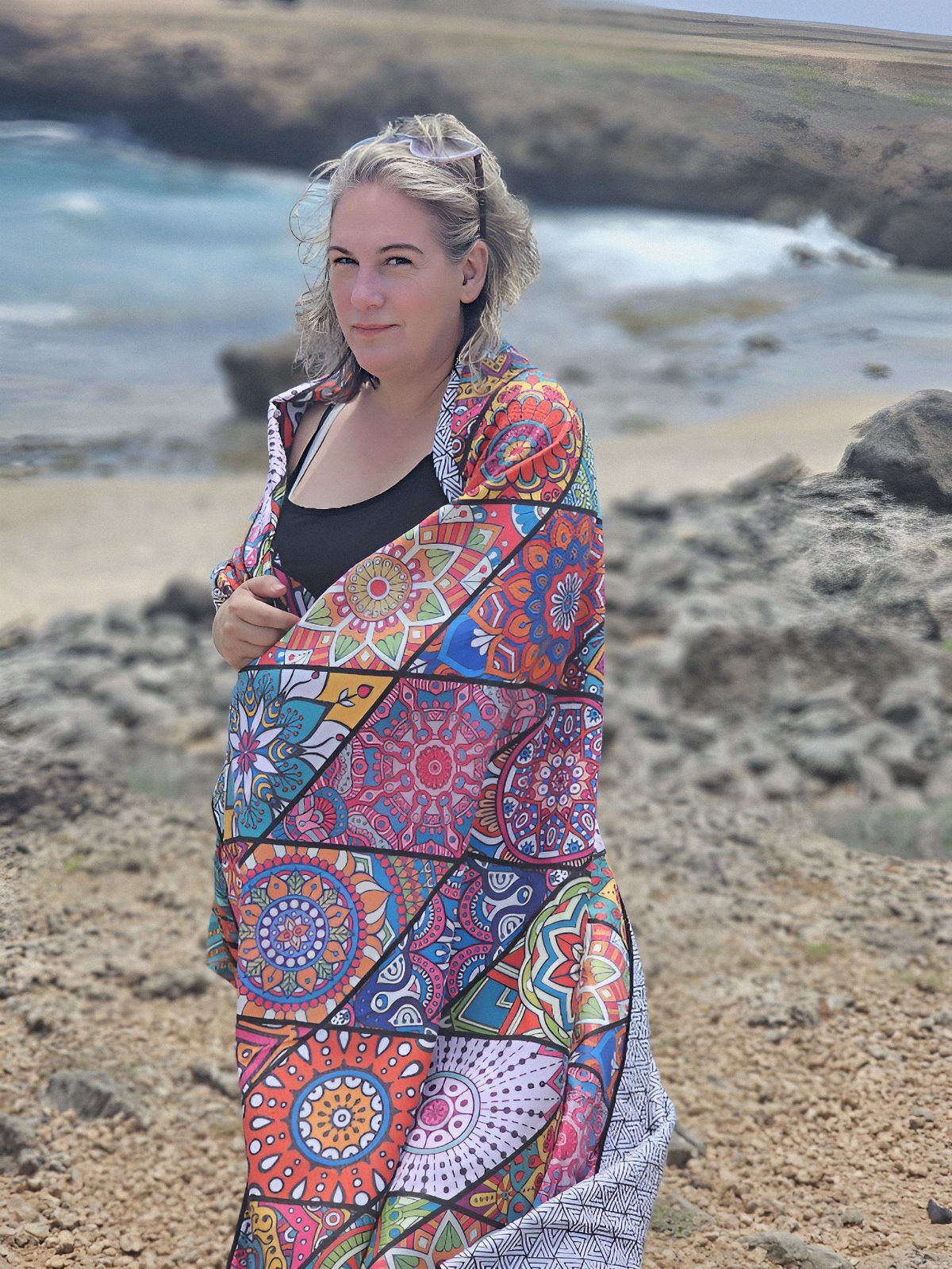 54-year-old travel blogger Kathy Brown stands on a cliff in Aruba, overlooking the Caribbean Sea. She wears a multi-colored beach blanket draped around her shoulders. The wind blows her grey hair in the breeze. The beach blanket moves in the breeze, creating a sense of peacefulness against the rugged terrain.