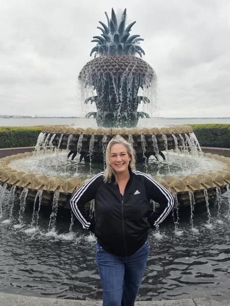 Kathy Brown, a stylish influencer over 50, poses in front of Charleston's iconic Pineapple fountain. She wears a black adidas zip-up jacket and jeans, embodying travel and lifestyle in this picturesque scene
