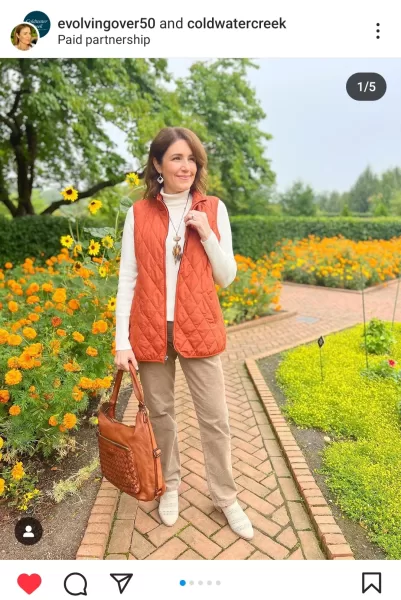 A stylish woman over 50, radiating classic beauty, elegantly dressed in a well-coordinated outfit. She wears an orange jacket and brown pants from Coldwater Creek, showcasing her partnership with fashion brands. Her brown hair frames her confident and fashionable appearance, as she highlights trending fashion for mature ladies in the USA on her Instagram feed.