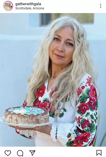 Gala, a stunning blonde woman in her 50s, wearing a lovely pink dress, proudly stands while holding a homemade designer cake. She exudes grace and elegance as she presents her delightful creation.