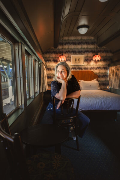 Chattanooga Midlife travel and lifestyle blogger Kathy Brown sits looking out a train car window at the outside courtyard of The Hotel Chalet.  In the background is a chic vintage king bed and luxurious decor.  Image Credit: Chattanooga travel and lifestyle blogger Kathy Brown gazing out the window of a train car.  Kathy is enjoying the view of The Hotel Chalet.  Behind this Chattanooga travel writer is a luxurious room with a king bed.  The overall aesthetic is vintage romance and boutique hotel luxury.  Image Credit: CHRISTIAN MICHELLE PHOTOGRAPHY